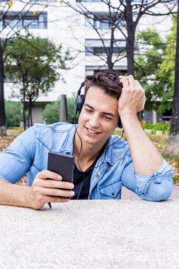 Kot pantolon giymiş neşeli genç adam, akıllı telefon ve kulaklık kullanırken, ağaçlarla kaplı bir şehir parkında otururken ve arka planda modern bir bina varken,.