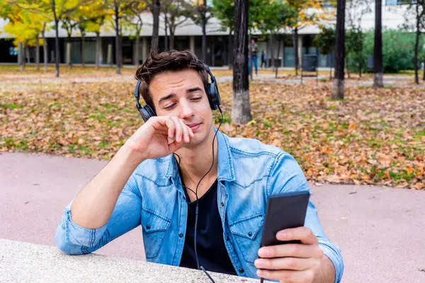 Young Male Denim Jacket Uses Headphones Smartphone Outdoors Leaning Bench Εικόνα Αρχείου