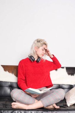 Sarı saçlı, kırmızı kazaklı ve ekose pantolonlu genç bir kadın, boynunda kulaklıklarla kanepede oturur, kafasını düşünceye daldırır okurken..
