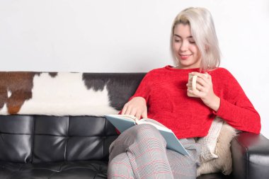 Sarı saçlı, kırmızı kazaklı ve ekose pantolonlu genç bir kadın, bir kanepede oturmuş, elinde kitap okuyor, sarı bir kupa tutuyor, gülümsüyor ve gevşiyor..