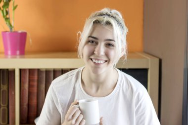 Genç sarışın kadın gülümsüyor ve elinde beyaz bir kupayla içeride oturuyor. Beyaz bir gömlek giyer ve bir kitaplığın ve turuncu bir duvarın önüne yerleştirilir..