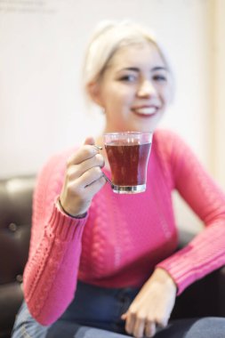 Pembe kazaklı genç bir kadın sıcak bir içeceği yudumluyor, ifadesi tatmin oluyor ve sıcak, aydınlık bir ev ortamında rahatlıyor, sıcak bir ortam yaratıyor.