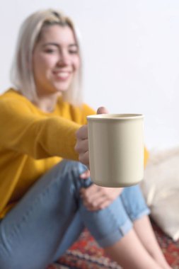 Kafkasyalı bir kadının bej bir kupa tutan eli, arka planda gülümseyen yüzü bulanık bir görüntüsü.