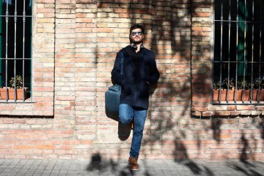 Donanma ceketli, kot pantolonlu ve botlu beyaz bir adam tuğla duvara yaslanıyor. Güneş gözlüğü ve kulaklık takıyor, çanta taşıyor. Kentsel, şık bir hava..