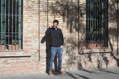 Siyah gömlek giyen beyaz bir adam telefonuyla konuşurken gülüyor, tuğla bir duvarın önünde duruyor. Parlak güneş ışığı neşe katar.