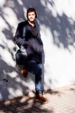 Donanma ceketi ve kot pantolon giyen, kulaklıklı beyaz bir adam, güneşli bir duvara yaslanıyor. Rahat bir şehir havası olan bir çanta taşıyan sıradan bir poz..