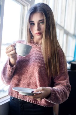 Kafkas kadın, uzun sarı saçlı, pencerenin yanında bir fincan çay tutuyor. Pembe bir kazak giyerek, düşünceli bir şekilde dışarı bakıyor..