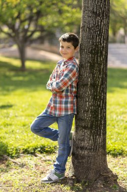 Kısa siyah saçlı, ekose gömlek ve kot giyen beyaz bir çocuk güneşli bir parkta ağaca yaslanmış, güvenle gülümsüyor..