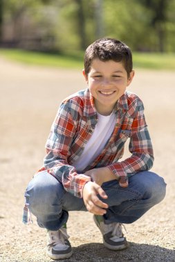 Kısa siyah saçlı, ekose gömlek, kot pantolon ve spor ayakkabı giyen beyaz bir çocuk, güneşli bir parktaki çakıl yolunda çömelmiş, meraklı görünüyor..