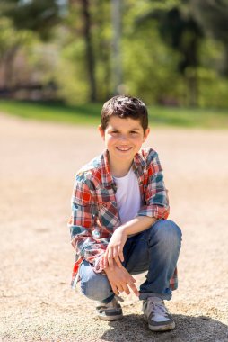 Kısa siyah saçlı, ekose gömlek, kot pantolon ve spor ayakkabı giyen beyaz bir çocuk, güneşli bir parktaki çakıl yolunda çömelmiş, meraklı görünüyor..