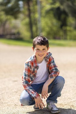 Kısa siyah saçlı, ekose gömlek, kot pantolon ve spor ayakkabı giyen beyaz bir çocuk, güneşli bir parktaki çakıl yolunda çömelmiş, meraklı görünüyor..
