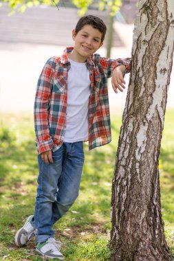 Kısa siyah saçlı, ekose gömlek ve kot giyen beyaz bir çocuk güneşli bir parkta ağaca yaslanmış, güvenle gülümsüyor..