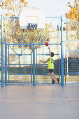 Neon sarısı formalı genç bir çocuk açık hava sahasında basket oynamaya kalkışıyor. Halka hasar görmüş ve altın yapraklı sonbahar ağaçları görülebiliyor..