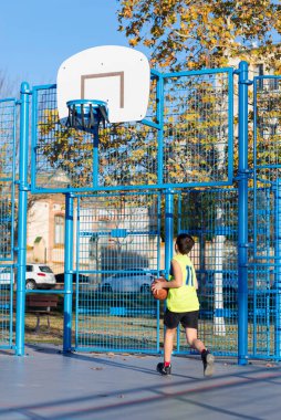 Neon sarısı formalı genç bir çocuk açık hava sahasında basket oynamaya kalkışıyor. Halka hasar görmüş ve altın yapraklı sonbahar ağaçları görülebiliyor..