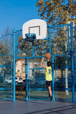 Neon sarısı formalı genç bir çocuk açık hava sahasında basket oynamaya kalkışıyor. Halka hasar görmüş ve altın yapraklı sonbahar ağaçları görülebiliyor..