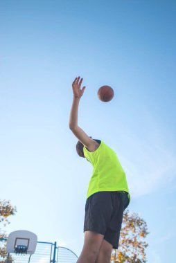 Neon sarısı formalı genç bir çocuk basket topunu yukarı fırlatıyor. Düşük açılı çekim açık mavi gökyüzüne ve arka planda bir mahkemeye karşı hareketi vurguluyor.