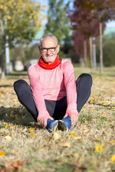 Parkta çimenlerde oturan, koşudan sonra esneyen, gözlüklü, pembe uzun kollu, siyah taytlı beyaz bir adam..