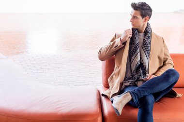 Modern deri koltukta oturan beyaz bir genç, deve ceketi, kot pantolon ve atkı giyiyor. Rahat ve kendinden emin bir şekilde yan tarafa bakıyor..