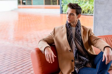 Modern deri koltukta oturan beyaz bir genç, deve ceketi, kot pantolon ve atkı giyiyor. Rahat ve kendinden emin bir şekilde yan tarafa bakıyor..