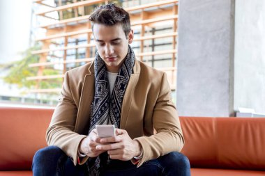 Açık havada deri bir koltukta oturan, akıllı telefon kullanarak gülümseyen beyaz bir genç adam. Deve kürkü ve eşarp giymiş, rahat ve şık bir duruşu var..