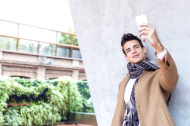Beyaz ceketli ve atkılı beyaz bir adam akıllı telefonuyla selfie çekiyor, beton bir duvarın yanında duruyor, gülümsüyor ve kendinden emin..