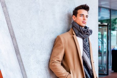Beyazlı genç adam bej ceket ve desenli eşarp giyiyor, beton duvarın önünde duruyor, düşünceli görünüyor. Şehir ortamında modaya uygun kış portresi.