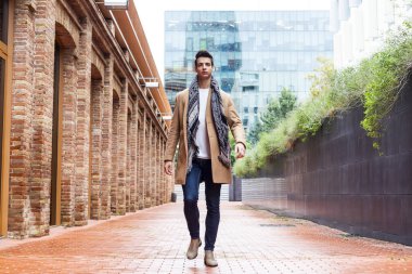 Tarz sahibi beyaz bir adam, eski tuğla duvarlar ve modern cam bina arasında güvenle yürüyor, deve kürkü ve eşarp giyiyor..