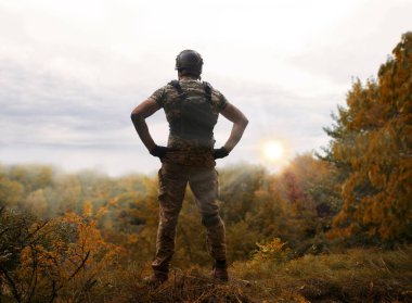 Alone tired young USA nato male guy stand rest hand hip waist look hope protect danger forest tree light white sky text space. Lone sad worry feel upset lost vet medic hero god faith worship concept clipart