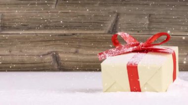 Karla kaplı kırmızı kurdeleli tek bir hediyeyle sakin bir hayat. Arka planda tahta tahta var. Düşen kar etkisi. Noel, tatil ve kış konsepti. Yüksek kalite 4k video.