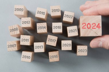 Geçen yılların gri sayılı tahta blok grubunun fotoğrafı ve bir tanesi ahşap yüzeye 2024 numaralı kırmızı damgalı bir insan eli tarafından tutuluyor. Yeni Yıl, Strateji, Gelecek ve Beklenen Konsept.