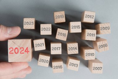 Geçen yılların gri sayılı tahta blok grubunun fotoğrafı ve bir tanesi ahşap yüzeye 2024 numaralı kırmızı damgalı bir insan eli tarafından tutuluyor. Yeni Yıl, Strateji, Gelecek ve Beklenen Konsept.