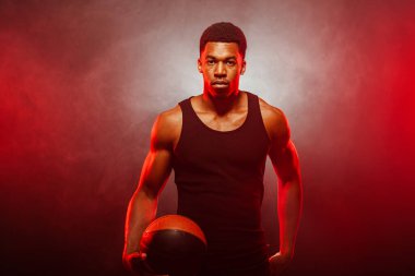 Basketbolcu tarafı kırmızı renkli, dumanlı arka planda topu tutuyor. Ciddi bir şekilde konsantre olmuş Afro-Amerikan erkeği..