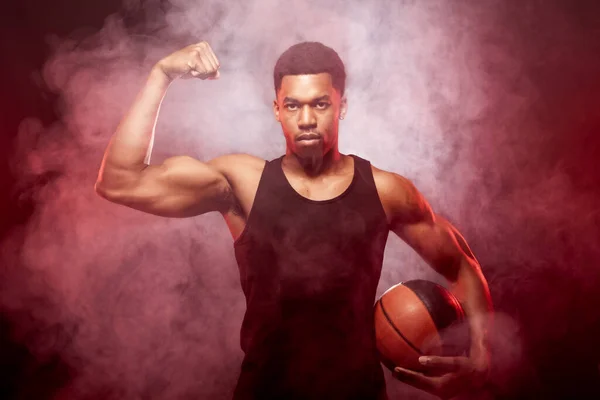 篮球运动员一边闪烁着红色的光芒 背后拿着一个烟雾弥漫的球 严重的非洲裔美国人表现出肌肉 — 图库照片