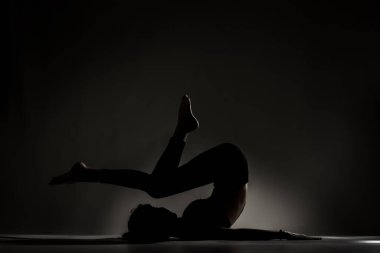 Fit balerin kız yalan söylüyor ve yoga pozları veriyor. Koyu arkaplana karşı yan aydınlatma silueti.