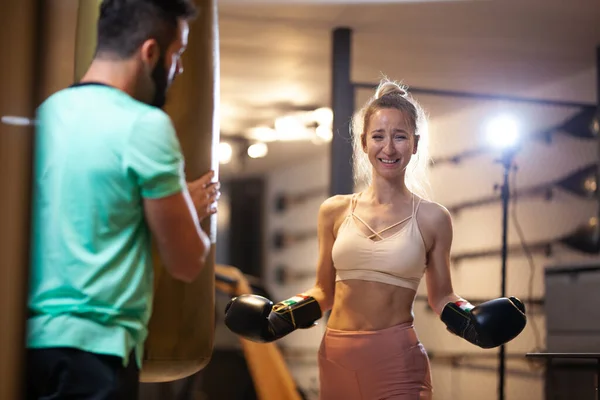 ボクシングの手袋を笑顔で女の子をフィット フィットネススタジオでバッグをパンチとキックボックスを練習女性クライアントと男性フィットネスインストラクター — ストック写真