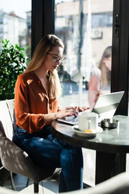 Bir kafe barında ya da parlak güneş ışığı olan bir restoranda genç bir iş kadını. Dizüstü bilgisayarda çalışan ve pencere kenarında çay içen serbest çalışan kız..