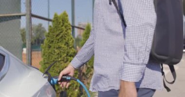 Elektrikli arabanın prizinden güç kablosunu çıkaran adam. Sırt çantası ve telefonu olan sıradan bir iş adamı..