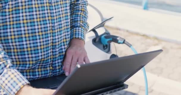 電気自動車の横にあるビジネスカジュアル衣装の男 彼の車が充電されている間ノートパソコンで働くビジネスマン 左カメラの動き — ストック動画