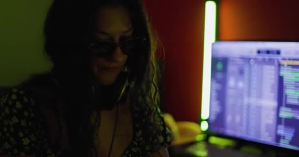 女性の音楽プロデューサーは オーディオ録音や監視機器とスタジオでキーボード楽器を演奏 ヘッドフォンとメガネの女の子 カラフルな点滅灯 — ストック動画