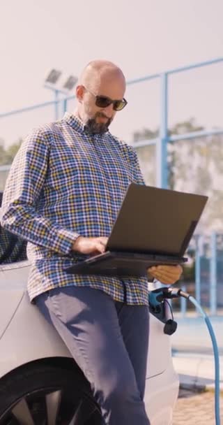 電気自動車の横にあるビジネスカジュアル衣装の男 彼の車が充電されている間ノートパソコンを使用してビジネスマン 垂直ビデオ — ストック動画
