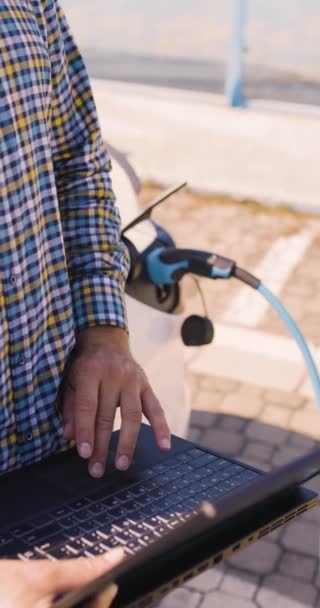 電気自動車の横にあるビジネスカジュアル衣装の男 彼の車が充電されている間ノートパソコンを使用してビジネスマン 垂直ビデオ — ストック動画