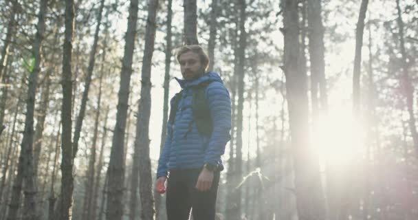 スポーツバックパックを着て森の中に立ってトレイルランナーの男の肖像画 日没の森の中の木を通して輝く太陽 スローモーションビデオ — ストック動画