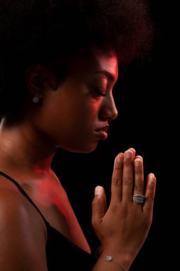Studio portrait of elegant african american lady with curly hair afro hairstyle against black background. Girl in black dress praying.