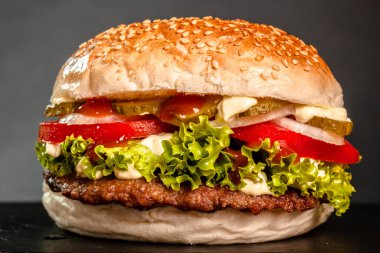 Hamburger with onions tomatoes cucumber and lettuce against dark gray background.