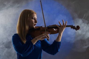 Beautiful blond girl playing violin. Female violinist against dark blue background with smoke.