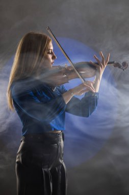 Beautiful blond girl playing violin. Female violinist against dark blue background with smoke.