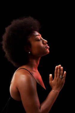 Studio portrait of elegant african american lady with curly hair afro hairstyle against black background. Girl in black dress praying.