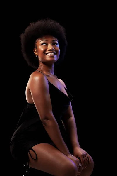 Studio portrait of elegant african american lady with curly hair afro hairstyle against black background. Happy girl smiling in black dress.