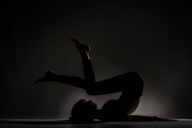 Fit balerin kız yalan söylüyor ve yoga pozları veriyor. Koyu arkaplana karşı yan aydınlatma silueti.