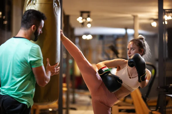 パンチングバッグを打つボクシンググローブで女の子をフィット フィットネススタジオでキックボックスを練習している女性のクライアントと男性フィットネスインストラクター — ストック写真